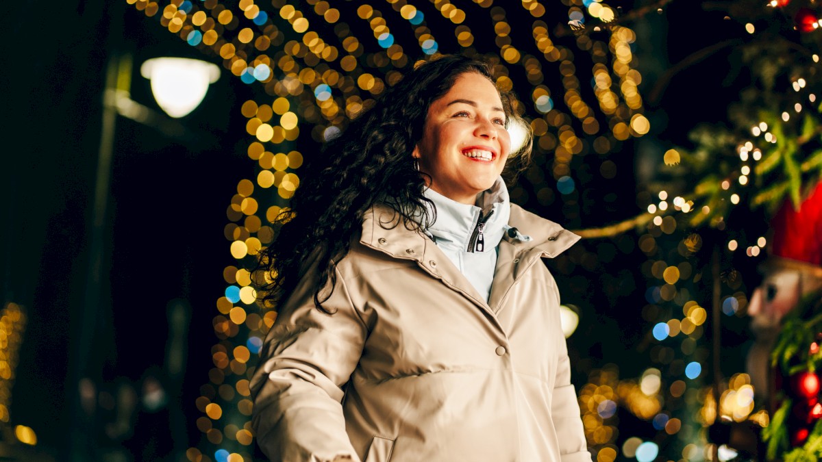 A joyful woman in a beige coat strolling at night among festive lights and decorations, smiling as holiday blooms glow around her.