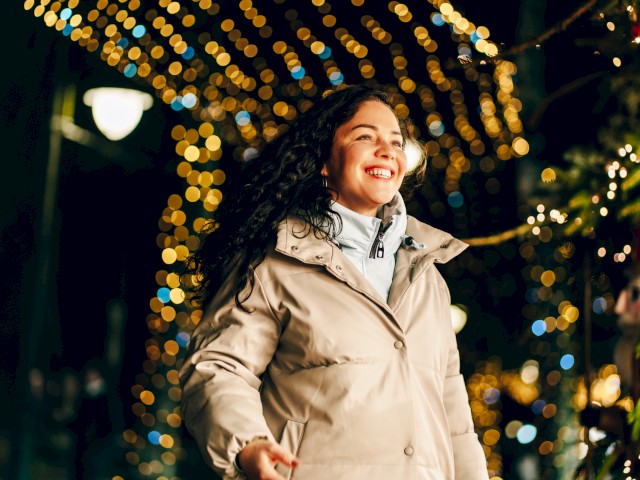 A smiling woman in a beige coat walks at night under twinkling holiday lights, festive and joyful.