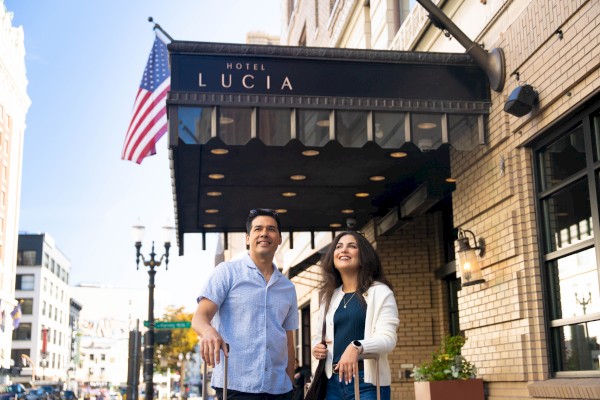 Two people stand outside a brick hotel with an American flag and a blue awning that reads “Hotel Lucía,” posing for a photo on a sunny street.