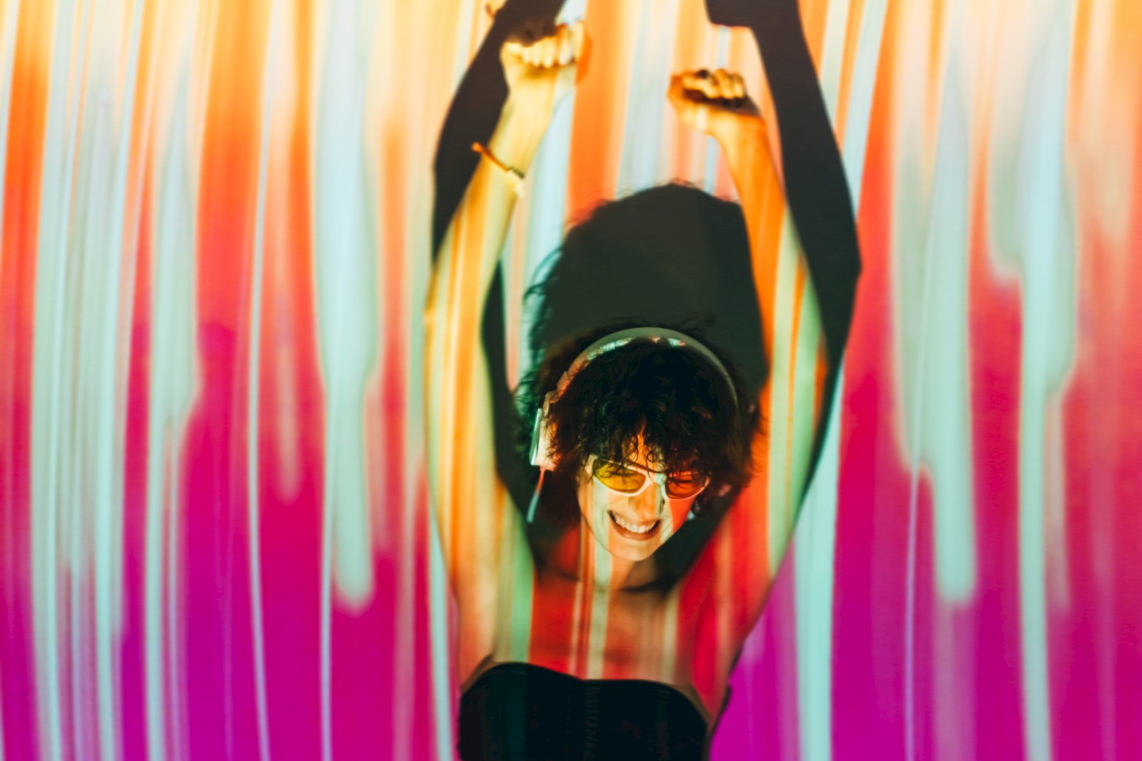 Colorful vertical stripes backdrop with a smiling person raising fists, wearing sunglasses and a black top, vibrant party vibes.