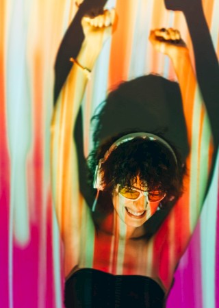 A person with short hair and sunglasses poses playfully against colorful vertical lights, arms raised in a fun, carefree vibe.