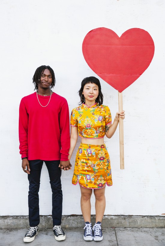 Two people stand side by side, the girl holding a large red heart on a stick, against a plain white wall.