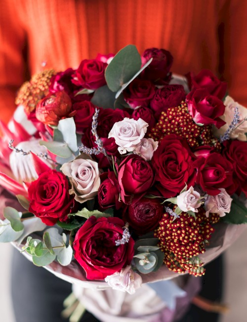 A bouquet of red, pink, and burgundy roses with eucalyptus and other flowers, held by a person in an orange top, wrapped in pink ribbon.