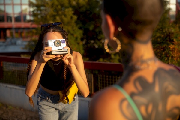 A woman with a camera aimed at another person, outdoors near a railing; the viewer’s back shows a tattooed shoulder and large hoop earrings.