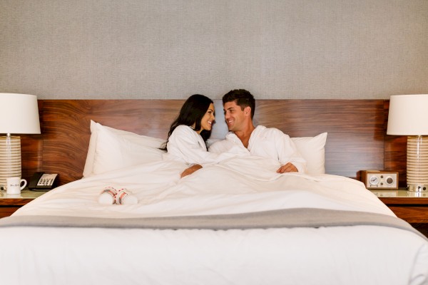 A couple sits up in bed, smiling at each other under white linens in a hotel room with two bedside lamps.