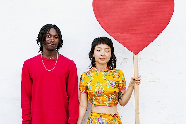 Two people stand side by side, holding hands, with the woman holding a large red heart on a stick, against a plain white wall.