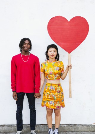 Two young people stand against a white wall, holding hands. The girl in a yellow floral dress holds a large red heart on a stick. End.