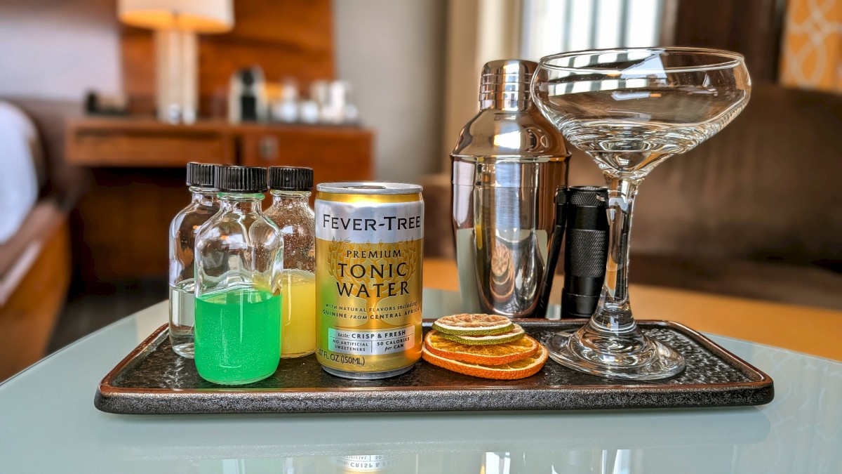 A hotel minibar tray with green liquid in a bottle, tonic water, a shot bottle, orange chips, cocktail glass, and shaker on a metal tray.