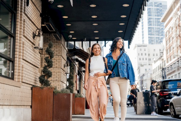 Two women with backpacks walk happily past the Hotel Lucia in an urban city setting, enjoying a sunny day.