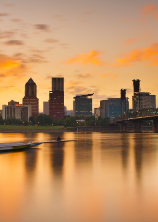 A city skyline at sunset over a calm river with boats and a bridge, surrounded by colorful clouds and reflections.