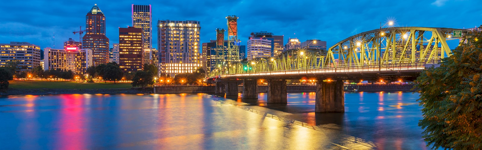 A vibrant city skyline at dusk with colorful lights reflecting on a river, featuring a bridge in the foreground and a cloudy sky.