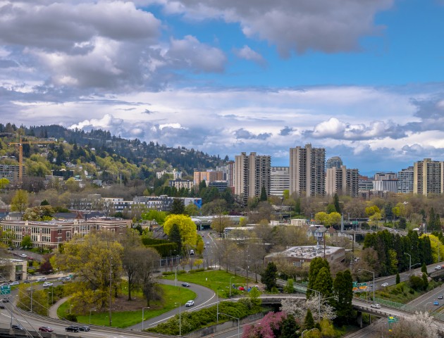 A cityscape with tall buildings, a hill covered in trees, and a partly cloudy sky.