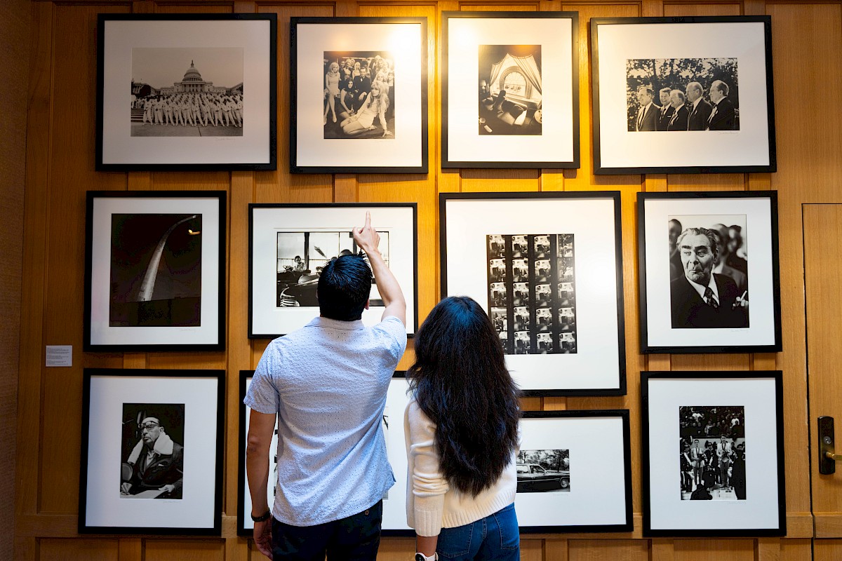 Two people are viewing black-and-white photographs displayed on a wooden wall in an art gallery or museum.