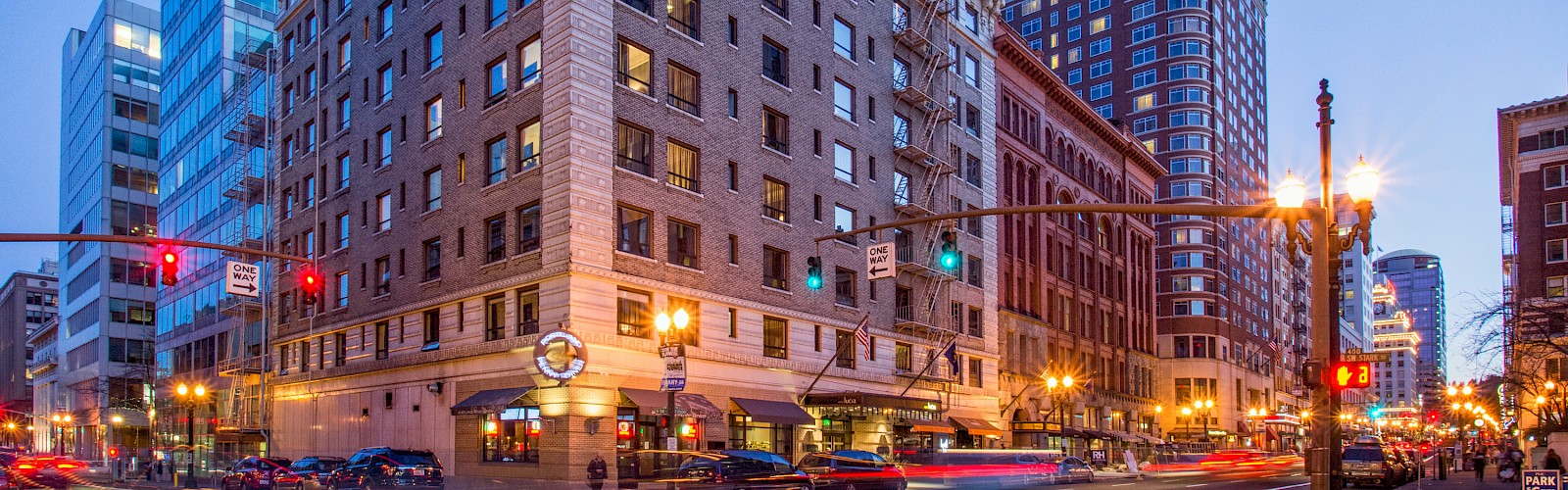 This is a city street scene showing tall buildings, cars, streetlights, traffic signals, and a mix of modern and historic architecture at dusk.