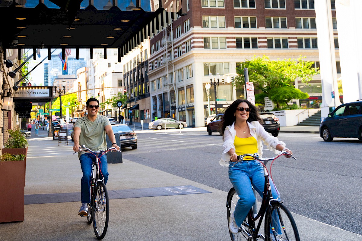 Two people are riding bikes on a city sidewalk with buildings, trees, and cars in the background on a sunny day.