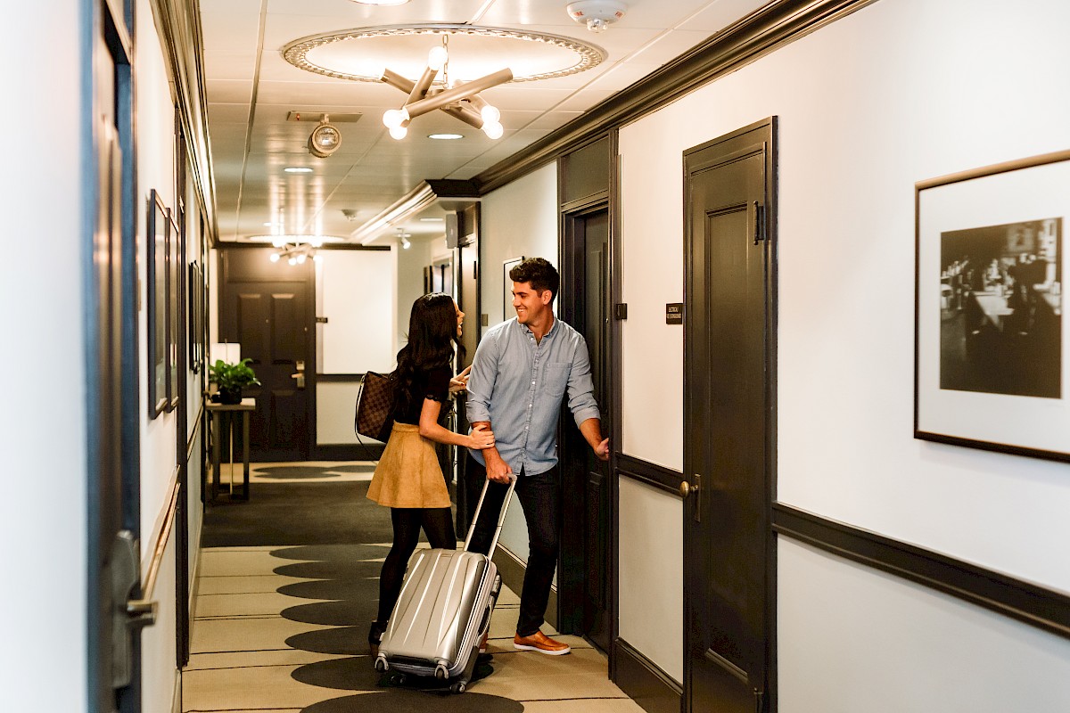 A couple with a suitcase smiles and chats in a hotel hallway.