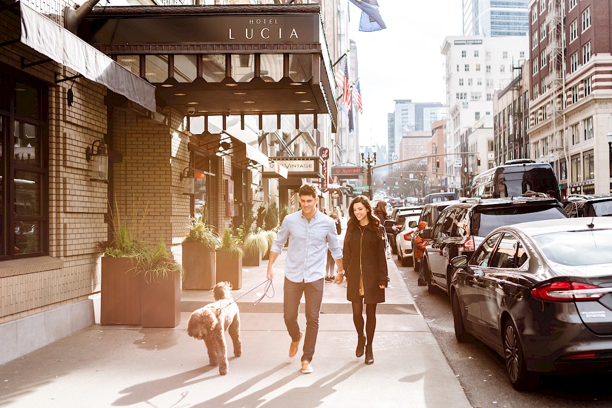 A young couple walks hand-in-hand with their dog on a busy city sidewalk lined with parked cars and tall buildings.