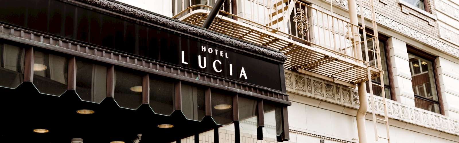 The image shows the Exteriors of Hotel Lucia, a building with a black marquee sign and orange awnings, featuring a staircase and a blue flag.