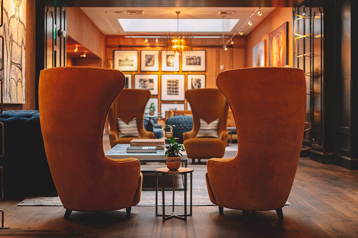 A cozy, stylish lounge with high-backed orange chairs, framed artworks on the wall, and a small table with a potted plant in the center.