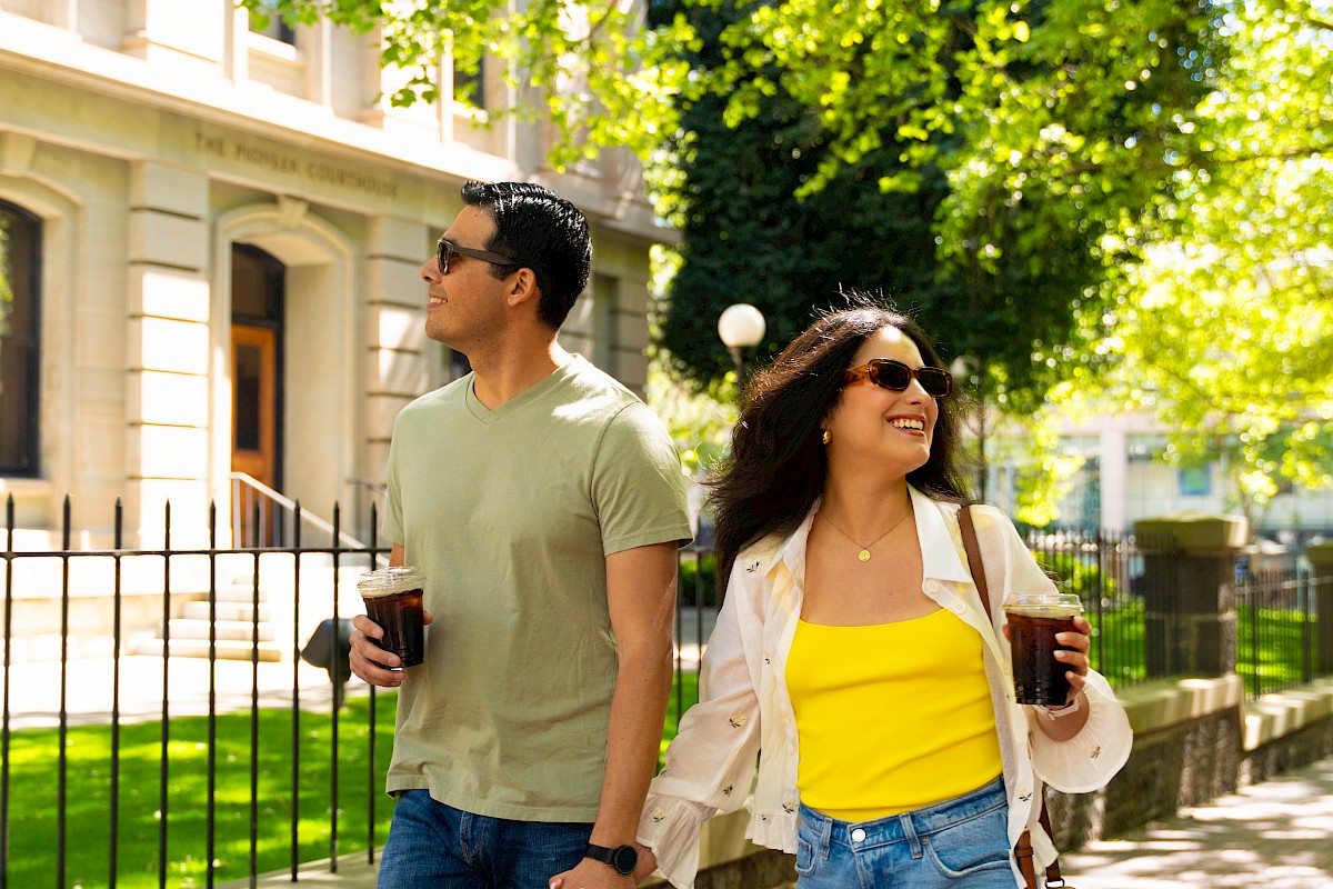 A happy couple walks hand in hand on a sunny city sidewalk, holding drinks, surrounded by vibrant green trees and historic buildings.