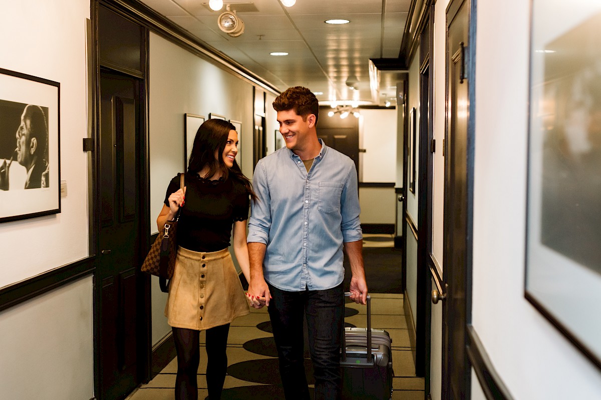 A happy couple walks hand-in-hand down a hotel corridor with luggage, smiling and enjoying each other's company.