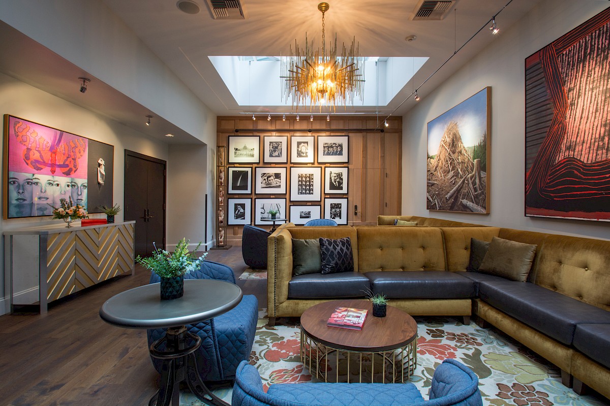 The image shows a stylish living room with colorful artwork, a mustard sofa, blue chairs, and a gallery wall of black-and-white photos.