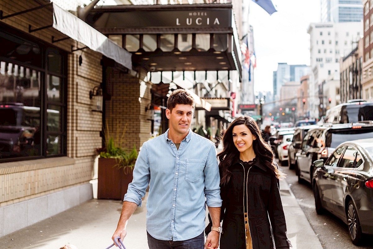 A young couple walks hand-in-hand on a city street with cars, tall buildings, and a sign for Hotel Lucia in the background.