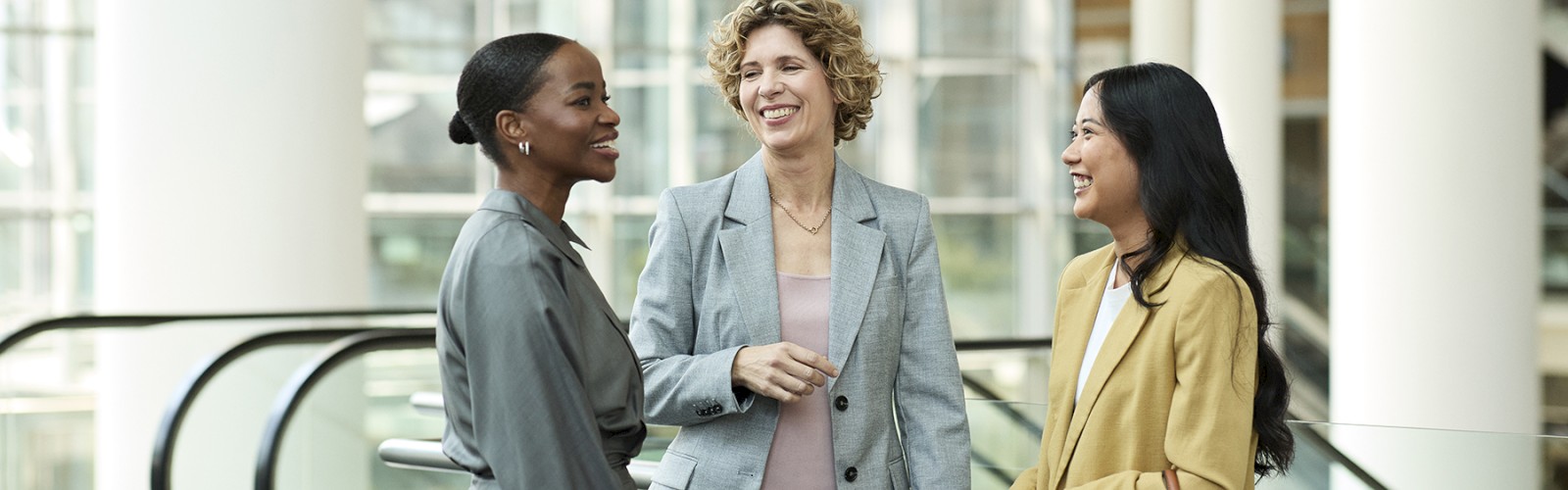 Three professional women are happily socializing in a modern office building lobby.