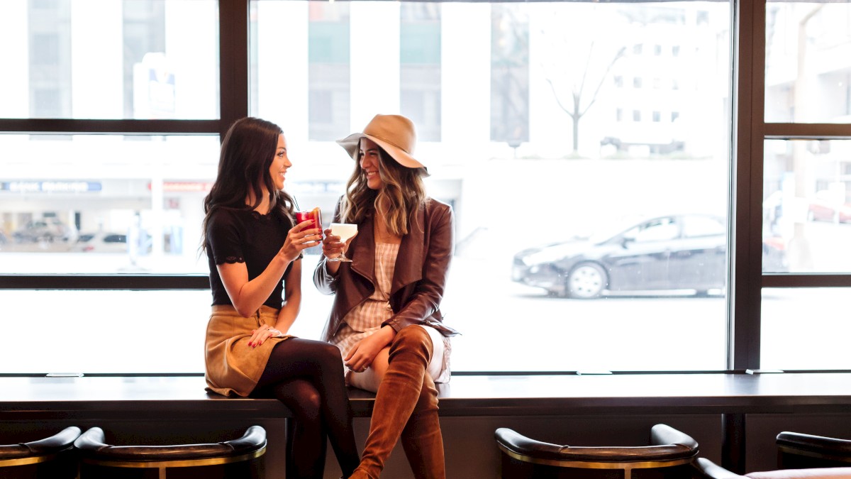 Two women sit by a large window in a cafe, laughing and toasting drinks, enjoying each other’s company on a bright day.