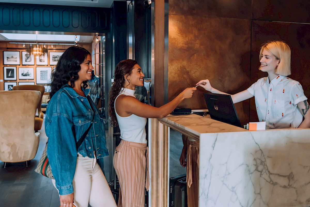 Two women check-in at a hotel reception desk, exchanging smiles while standing in line.