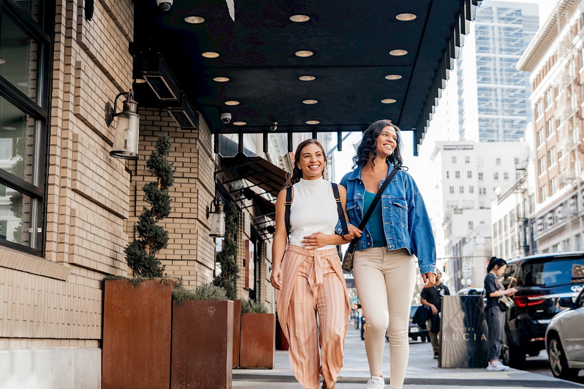Two women with backpacks walking and smiling outside Hotel Lucia on a busy city street.