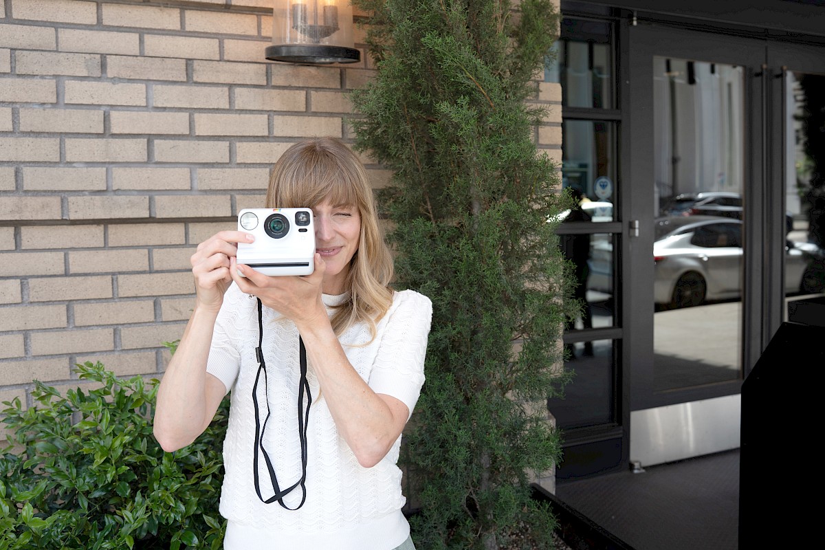 A woman with light brown hair is smiling and taking a picture with a Polaroid camera outside a building.