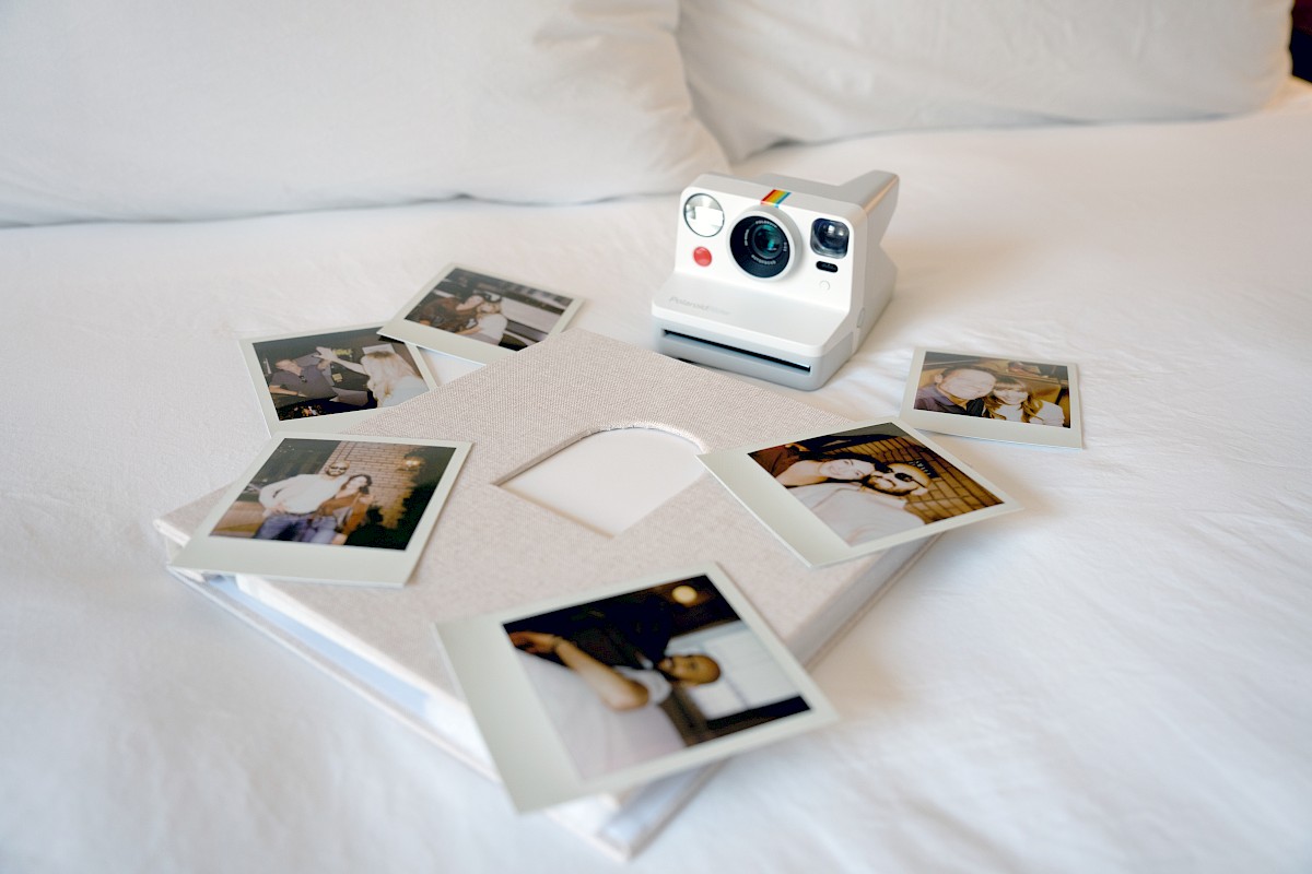 A Polaroid camera and printed photos are spread out on a white bed, with some pictures of people and a white book or box beneath them.