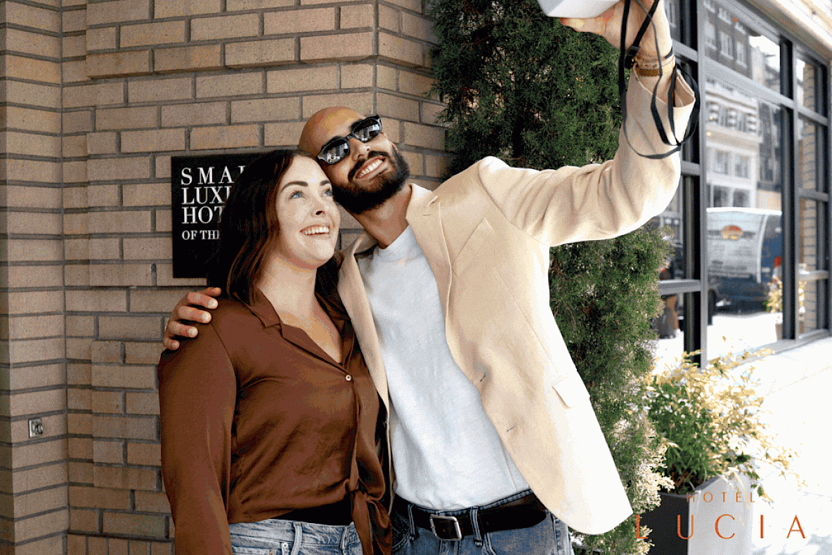 A smiling couple takes a selfie outside a hotel named Lucia, with one person wearing sunglasses and a cream jacket.