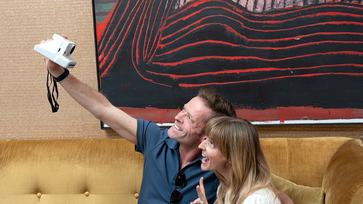 A couple happily takes a selfie together on a yellow couch in an art-filled room.