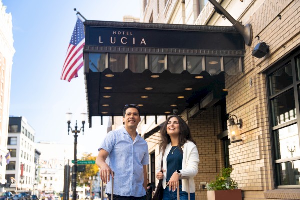 A man and a woman stand outside Hotel Lucia, smiling and looking up on a sunny city street.
