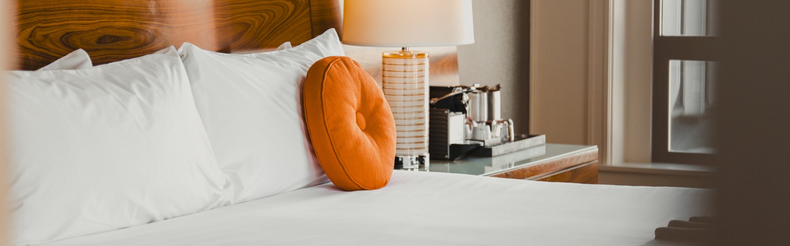 A neatly made hotel bed with white linens, a round orange pillow, a bedside lamp, and a window providing natural light.